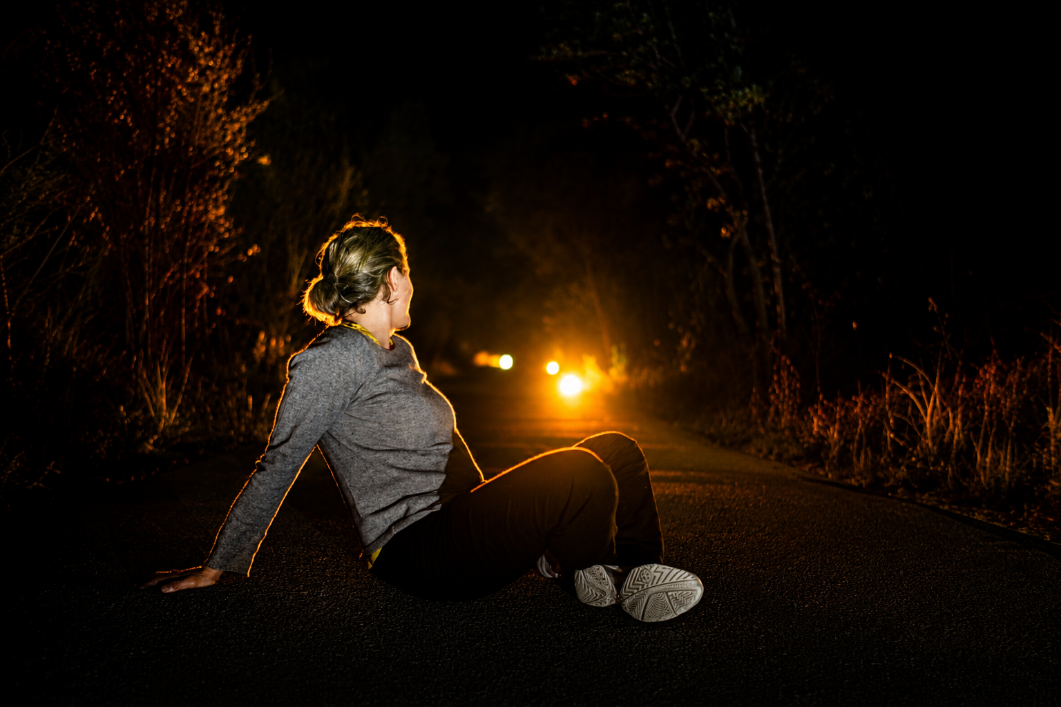 A woman sitting on a quiet road at night, lit by warm headlights